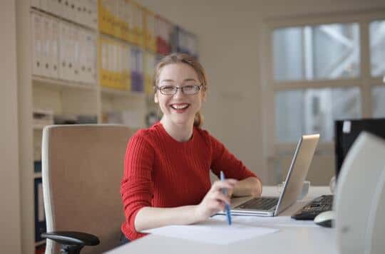 Smiling woman in red sweater working at desk with laptop and papers, holding a pen in a bright office with shelves behind her.